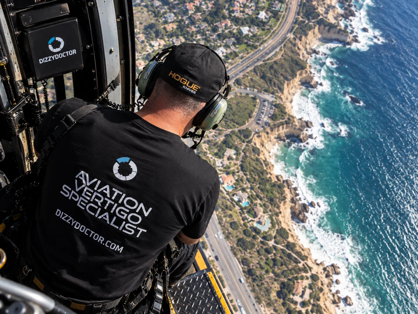 Dr. Ian Purcell seated in an open helicopter doorway above the coastline, wearing an Aviation Vertigo Specialist shirt, with DizzyDoctor recording equipment mounted beside him